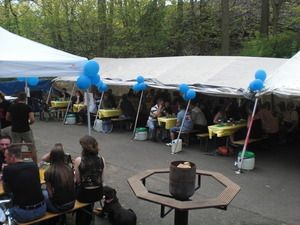 Viele Menschen die in einem Zelt sitzen - die Seitenwände sind hochgeklappt. Im Vordergrund sieht man einen Feuertisch. Dieser brennt aber nicht. Es ist Tag.